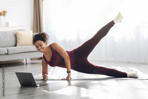 Sporty african american woman exercising at home, using laptop