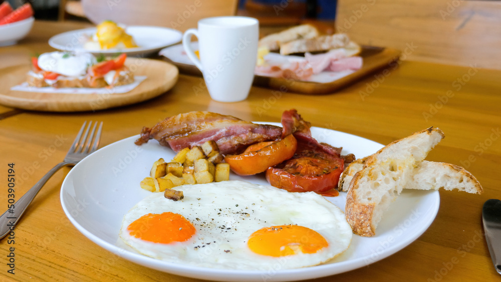 American breakfast with sunny side fried eggs, bacon, tomatoes, bread toasts, baked potatoes and coffee cup on wooden table in restaurant. Delicious served lunch or brunch food set meals in cafe. American breakfast with sunny side fried eggs, bacon, tomatoes, bread toasts, baked potatoes and coffee cup on wooden table in restaurant. Delicious served lunch or brunch food set meals in cafe.