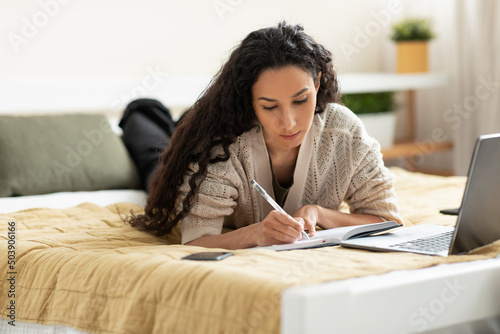 Woman lying on bed, using computer and writing in notebook photo