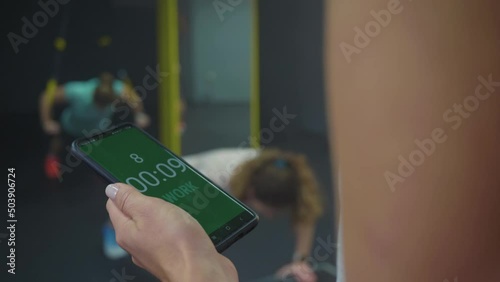 Caucasian female hand holding a smartphone with tabata timer app on a screen, measuring time intervals of tabata workout participants in the background, close up shot. photo