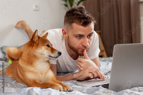 young man uses laptop while lying next to shiba inu dog 