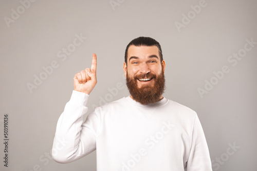 Cheerful bearded man is pointing up and smiling because he has got an idea. Studio shot over grey background.
