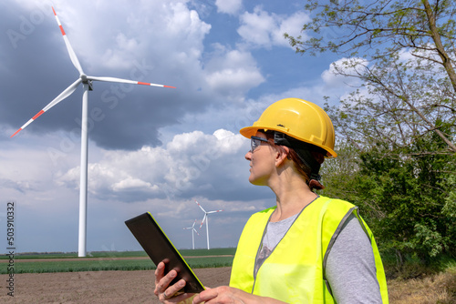 A female engineer standing on a field, looking at windmills on a wind farm, and making notes on a digital tablet. Green renewable energy concept.