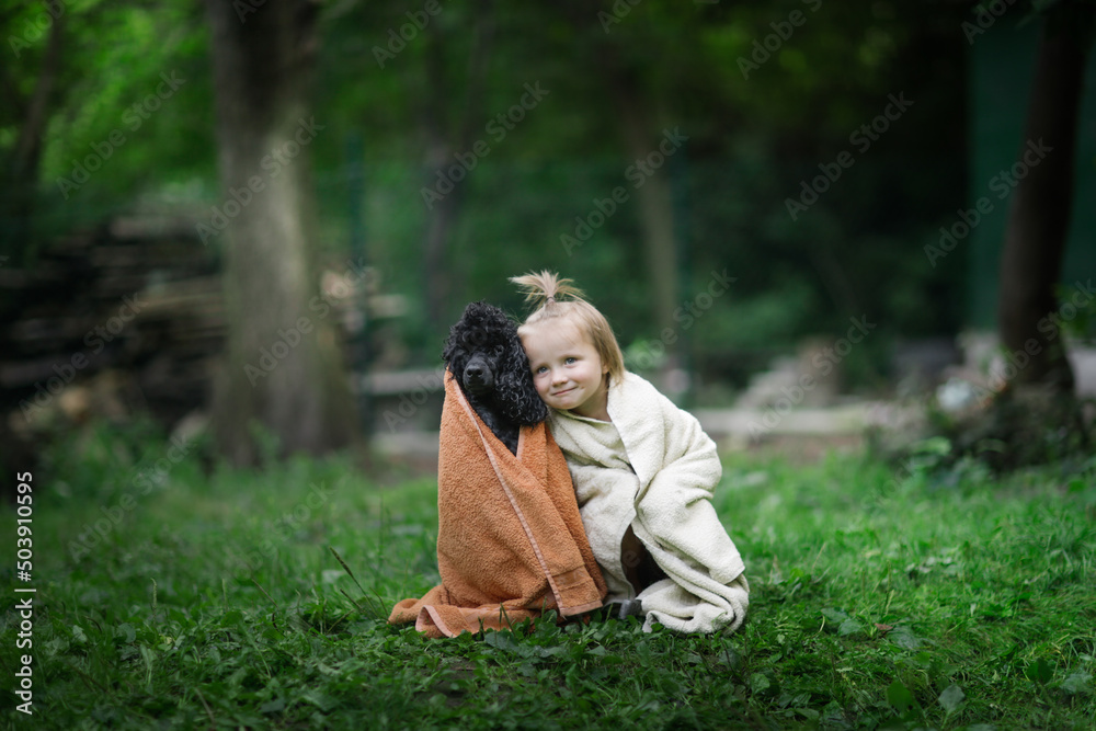 Cute baby toddler in towels with pet. Child and black poodle in a towel after swimming in the backyard in summer