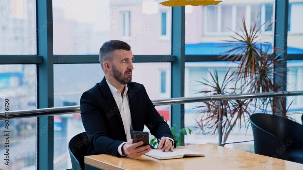 Businessman in suit sitting at desk holding a phone in one hand and pen in another. Man looks aside and talk strictly to someone. Businessman in suit sitting at desk holding a phone in one hand and pen in another. Man looks aside and talk strictly to someone.