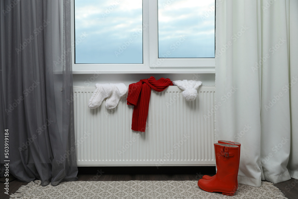 Obraz premium Heating radiator with hat, socks, scarf and rubber boots near window indoors