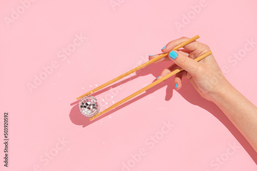 Creative layout with woman hand holding chopsticks and disco ball decoration on pastel pink background. 80s or 90s retro fashion aesthetic party concept. Minimal romantic food idea. photo