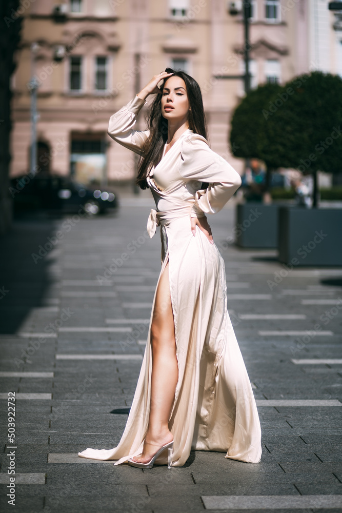 Sensual brunette beautiful woman luxury model posing in fluttering champagne color long dress on the street. Sensual brunette beautiful woman luxury model posing in fluttering champagne color long dress on the street.