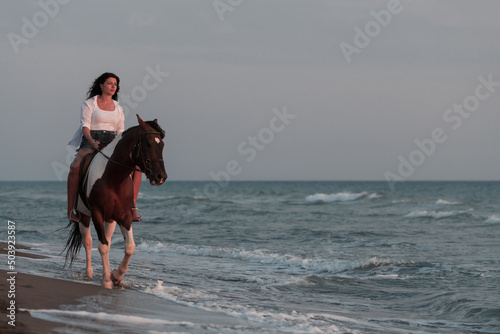 Foto Woman in summer clothes enjoys riding a horse on a beautiful sandy beach at sunset