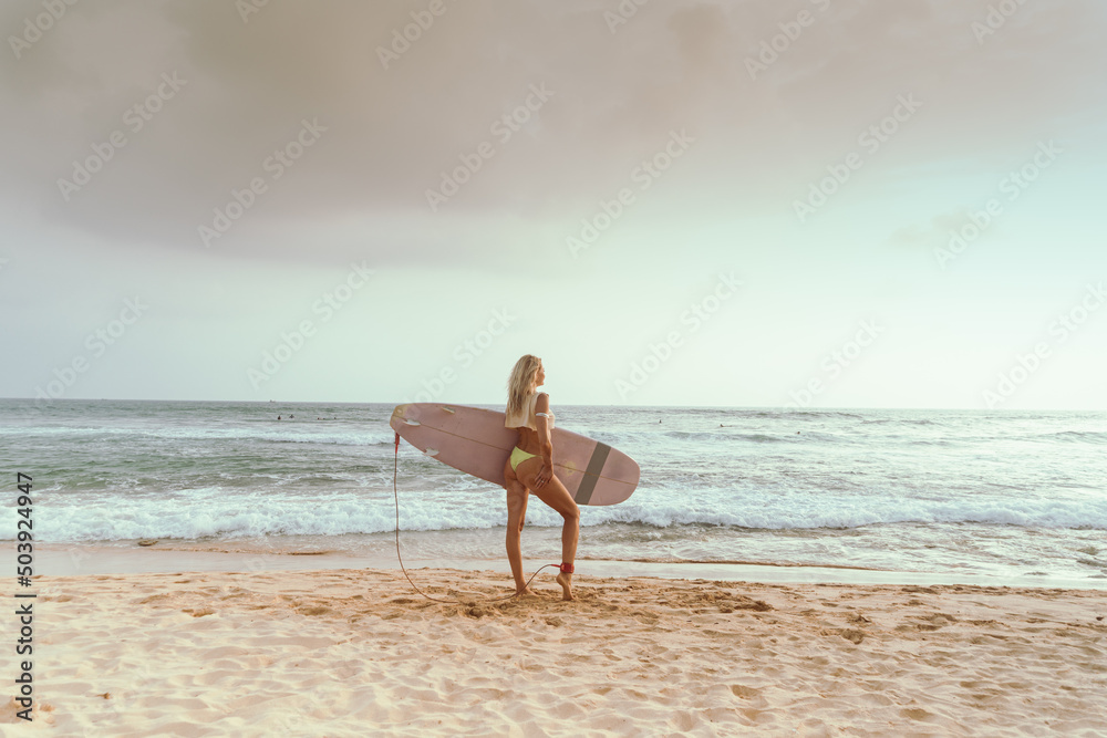 A girl surfer with a surfboard, in a swimsuit, on the beach against the ocean.
