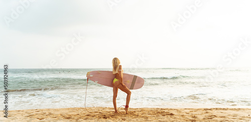 A girl surfer with a surfboard, in a swimsuit, on the beach against the ocean.