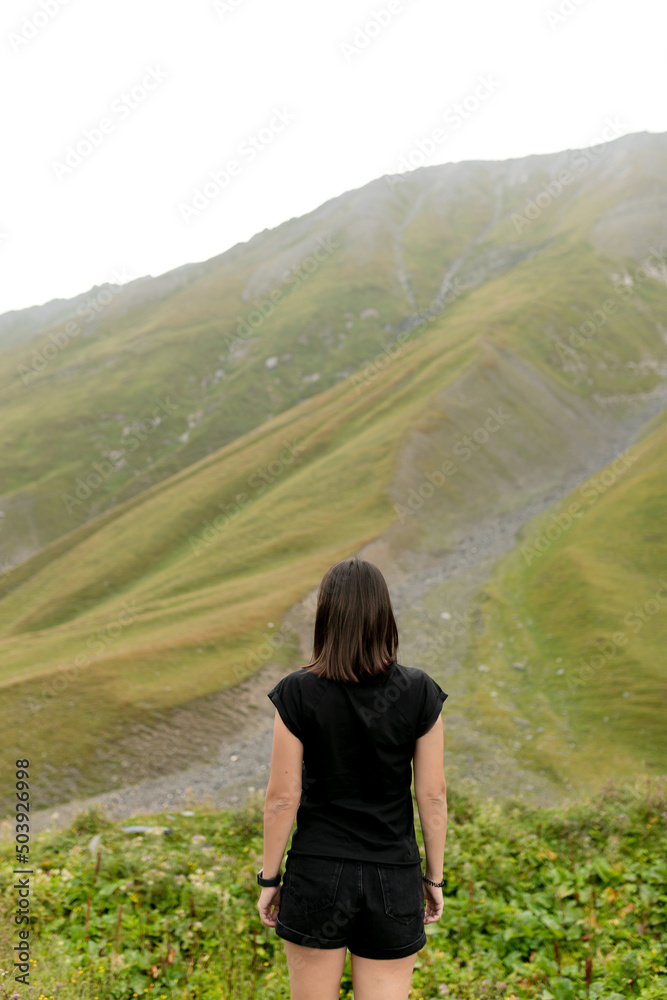 Portrait from back of lovely cute brunette girl wearing black t-shirt and shots standing among beautiful green mountains 