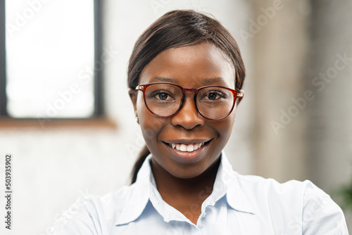 Close-up portrait of charming positive African-American business woman wearing stylish eyeglasses and formal shirt. Confident female office employee looks at the camera.