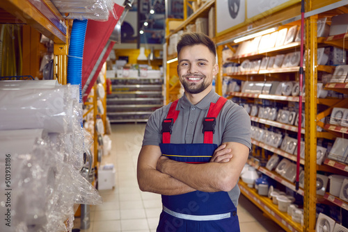 Confident friendly man working in warehouse in hypermarket of building materials. Smiling young male worker in coveralls standing with folded arms in one of aisles between shelves with goods. photo