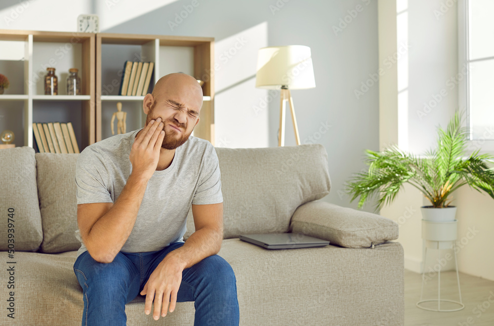 Young man with painful expression holds his cheek suffering from severe toothache. Upset unhappy Caucasian man sits on sofa at home in living room and suffers from worst pain. Dental treatment concept Young man with painful expression holds his cheek suffering from severe toothache. Upset unhappy Caucasian man sits on sofa at home in living room and suffers from worst pain. Dental treatment concept