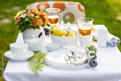 Elegant table setting with white dishes on a white linen tablecloth. Ripe fruits, drinks in crystal glasses. Summer holiday table set up. Table set for outdoor eating in the garden. Soft focus