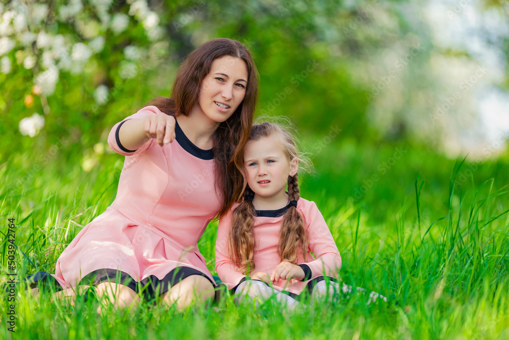 mother and daughter sit in the green grass against the backdrop of blooming apple trees mother and daughter sit in the green grass against the backdrop of blooming apple trees