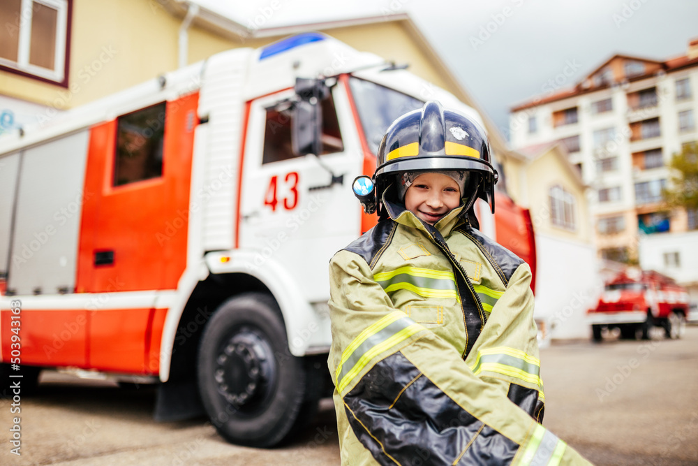 Obraz premium Child, cute boy, dressed in fire fighers cloths in a fire station with fire truck, childs dreamed profession