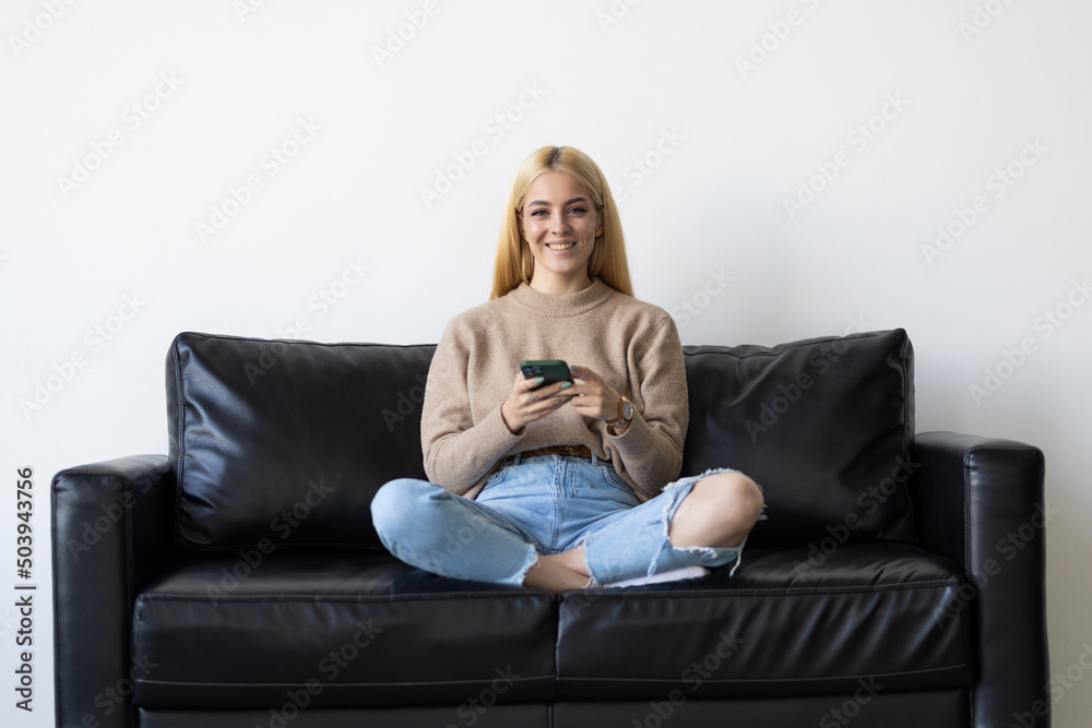 Obraz premium Happy young woman using mobile phone while sitting a couch at home with laptop computer