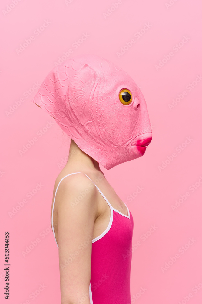 A woman in a pink fish head mask stands in profile against a pink  background and looks into the camera with one yellow eye, a crazy  conceptual Halloween costume Stock Photo