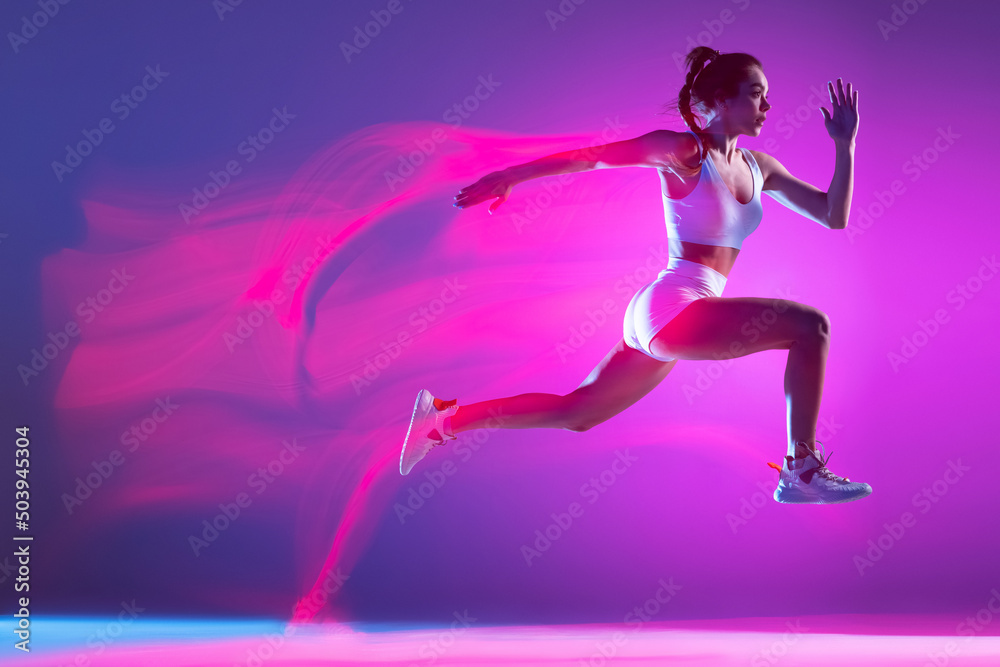 Professional female athlete, runner training isolated on blue studio background in mixed pink neon light. Healthy lifestyle, sport, motion and action concept.