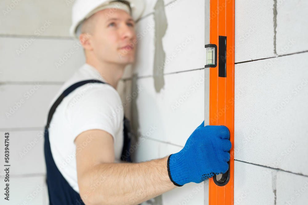 Beautiful young architect or builder in a white hard hat checks the level of the wall with the level tool. Beautiful young architect or builder in a white hard hat checks the level of the wall with the level tool.