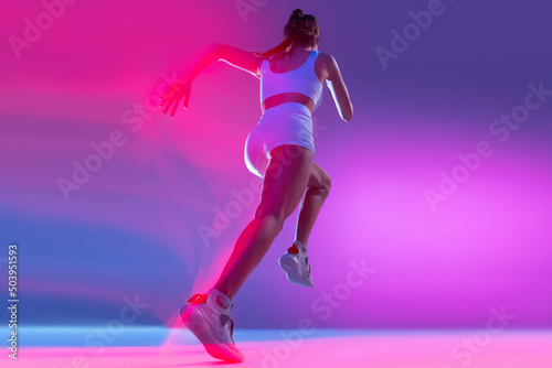 Professional female athlete, runner training isolated on blue studio background in mixed pink neon light. Healthy lifestyle, sport, motion and action concept. © master1305