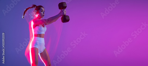 Flyer. Young sportive girl training with weights isolated on gradient blue-pink studio background in neon light. Sport, action, fitness, youth concept.