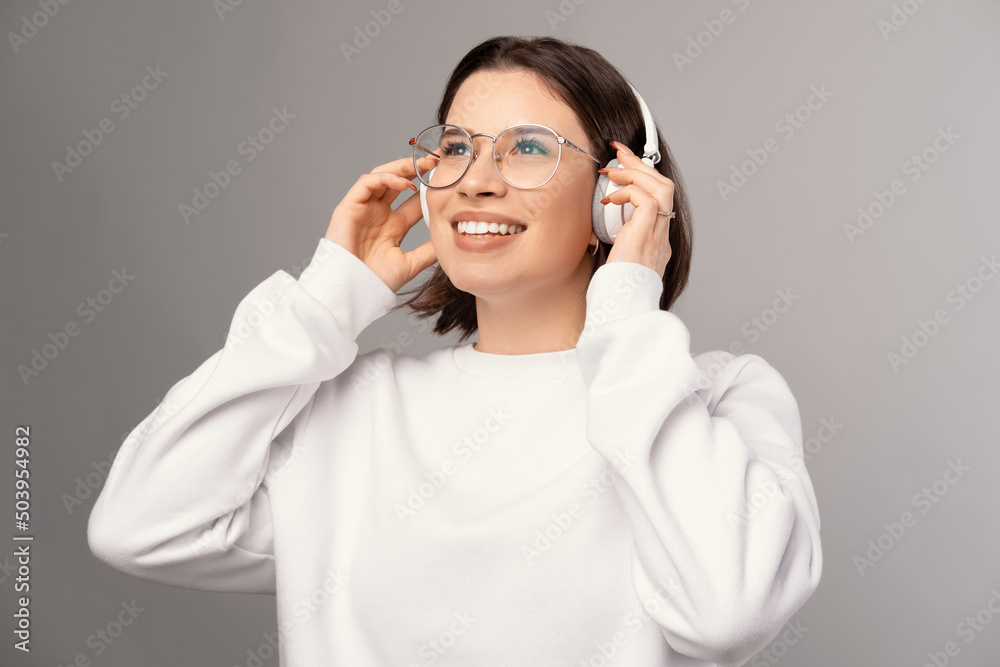 Young smiling woman wearing glasses and headphones enjoys music. Studio shot over grey background. Young smiling woman wearing glasses and headphones enjoys music. Studio shot over grey background.