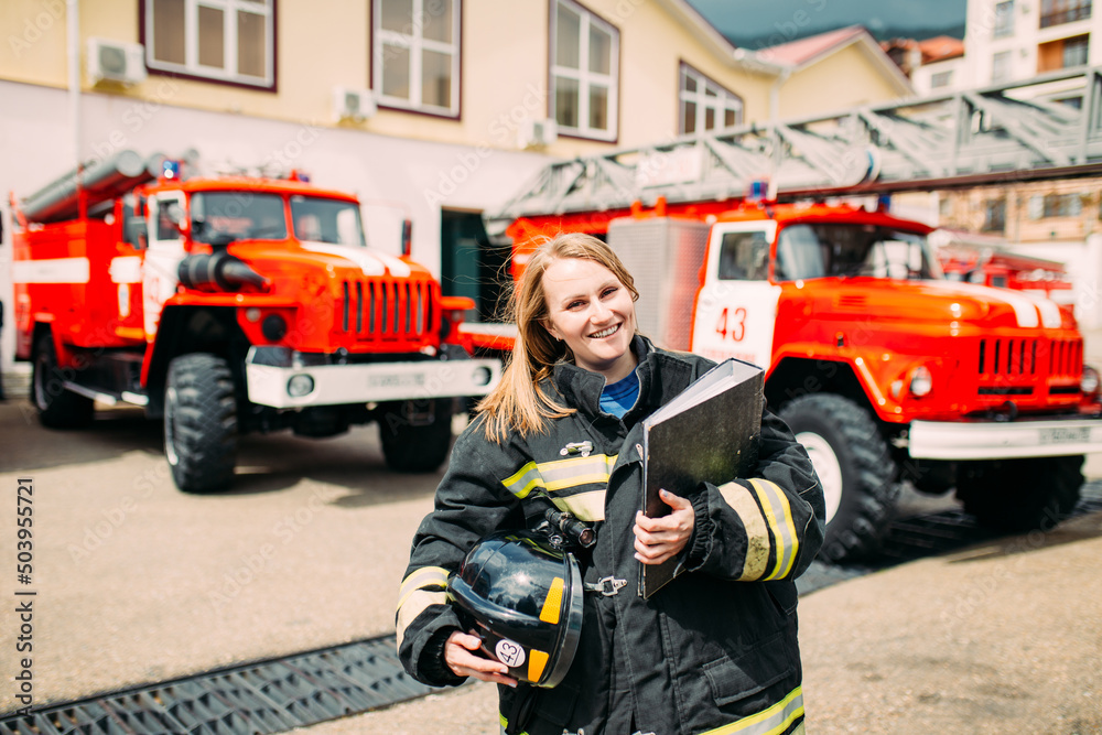 Female firefighter in in a protective suit with documents standing ain the background of a fire engines. Fire station Female firefighter in in a protective suit with documents standing ain the background of a fire engines. Fire station