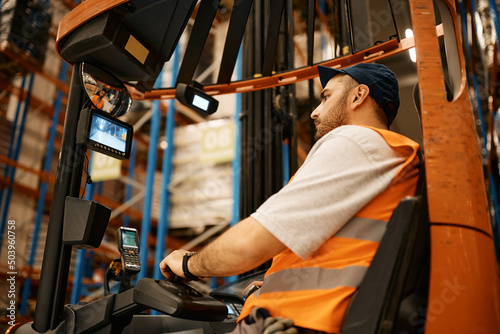 Young man driving forklift while working at storage compartment of distribution warehouse.