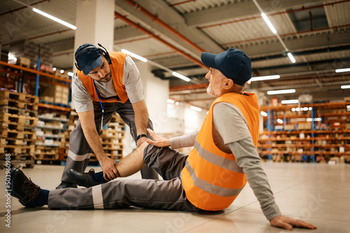 Young worker assists his colleague with leg injury while working at distribution warehouse.