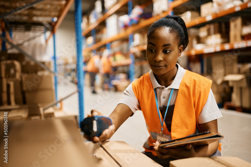 Black female worker using digital tablet and scanning labels on boxes while working in warehouse. photo