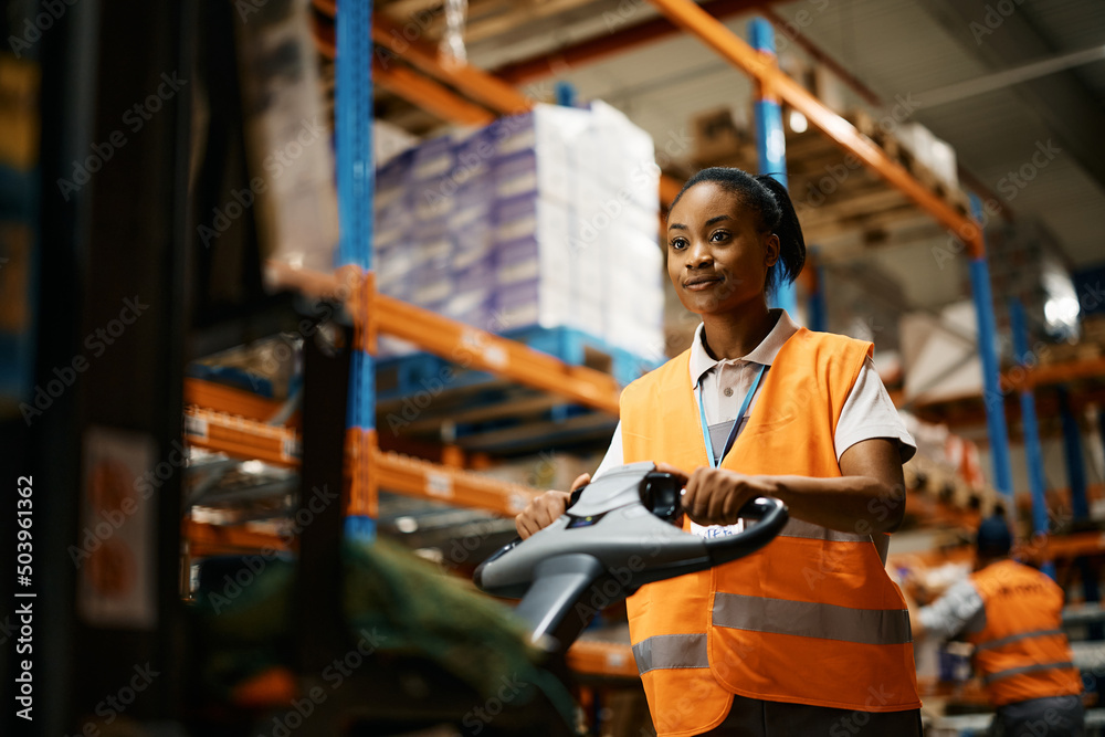 Black female worker pushing cargo on pallet jack while working at distribution warehouse.