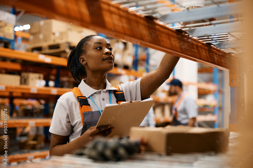 Black female worker checking inventory on shelves of distribution warehouse. - obrazy, fototapety, plakaty 