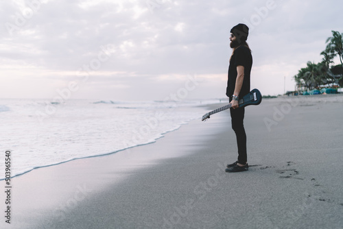 Side view of talented performer musician with professional Hawaiian guitar with six string creating sounds on leisure, stylish rock'n'roll man with ukulele enjoying music hobby for recreating at beach