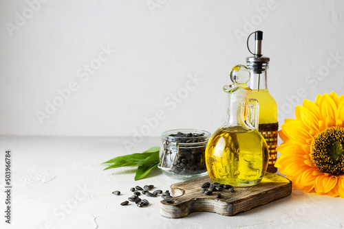Transparent jug with sunflower oil on a dark wooden plank. A bottle of butter and seeds in a glass jar in the background. Light background, space for text
