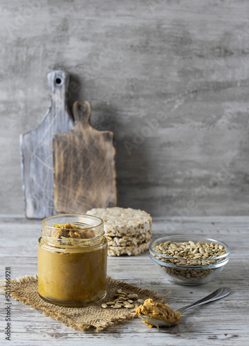 A glass jar with sunflower seed oil on a light wooden table. Crispy bread in the background. Seeds in a transparent vase on the table. Light background