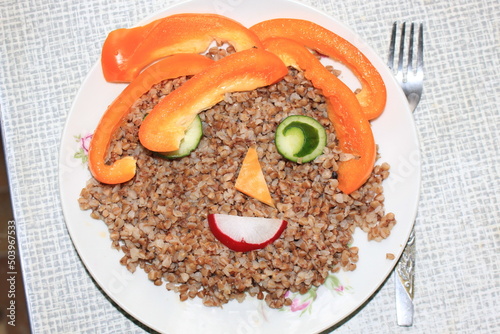 Breakfast for a child is decorated in the form of a funny face in a plate. Buckwheat, pepper, cucumber, cheese, sausage are good for the body