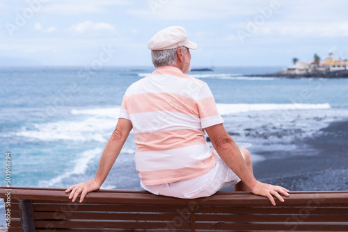 Rear view of senior man with beard and hat sitting outdoors at sea looking at horizon enjoying freedom and vacation. Active caucasian elderly man relaxing on a sunny day close to the beach © luciano