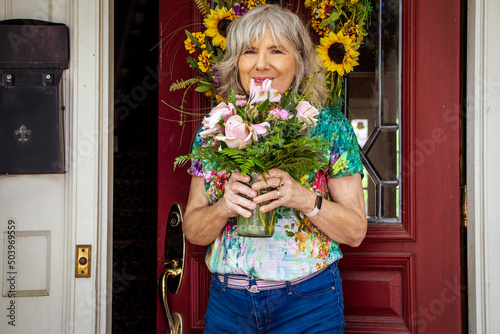 Older silver haired woman in jeans and floral tee-shirt  smiling at residence entrance door with leaded window and sunflower wreath holding just delivered pink flowers in vase