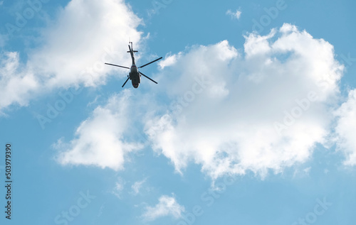 High, silhouette of helicopter flying high against blue sky with white clouds. Bottom view.
