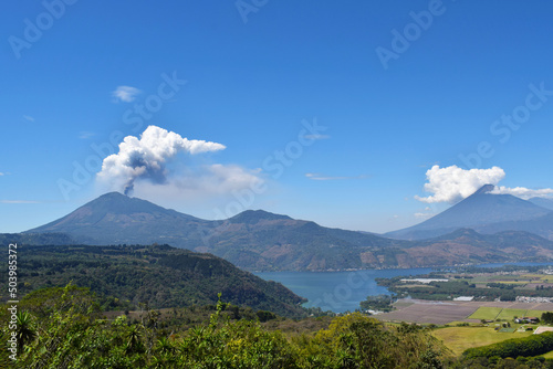 Panor  mica del volc  n Pacaya haciendo erupci  n al lado izquierdo  al lado derecho el volc  n de agua y en la parte de abajo el lago de Amatitl  n en Guatemala.