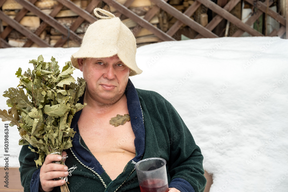 A man came out of the steam room with an oak broom, sits against the background of white snow, drinks a drink