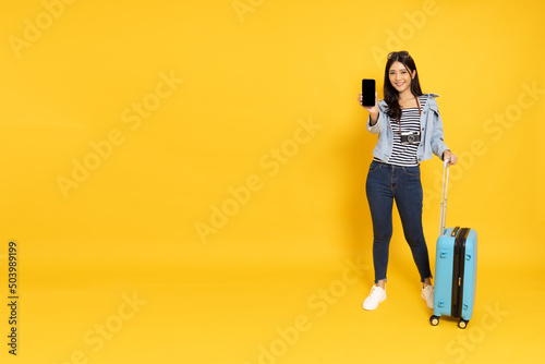 Happy Asian woman traveler standing and showing screen of smartphone isolated on yellow background, Tourist girl having cheerful holiday trip concept, Full body composition