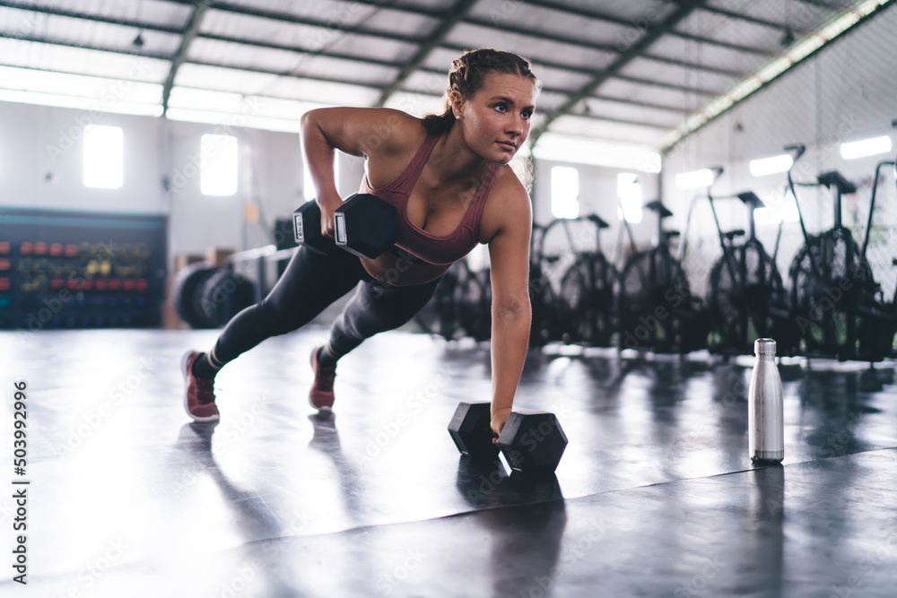 Caucasian female athlete in sportswear making push ups exercises during slimming workout training in gym, strong woman with dumbbells standing in hard plank on one hand practice intensive crossfit