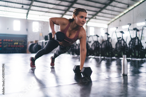 Caucasian female athlete in sportswear making push ups exercises during slimming workout training in gym, strong woman with dumbbells standing in hard plank on one hand practice intensive crossfit