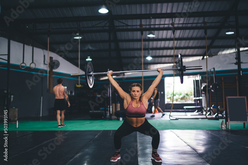 Portrait of fit young woman lifting barbell during hard bodybuilding workout in sportive studio gym, Caucaisan female athlete in tracksuit looking at camera while weightlifting and exercising