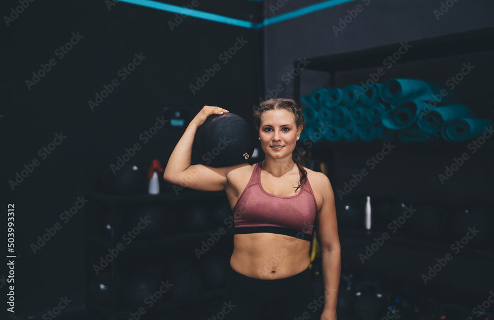Obraz premium Half length portrait of young Caucaisan female holding sportive ball and looking at camera during workout day for physical recreation, motivated athlete in tracksuit using gym equipment and posing