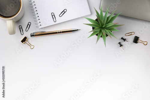 Working place with closed grey laptop, white cup of coffee, white notebook and paper clips on it, gold colored pen, black and gold colored paper clips and a plant in a pot. Place for text